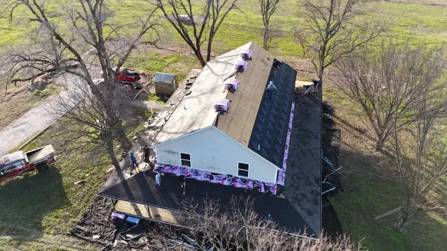 Aerial view of a roof replacement with underlayment going on, surrounded by farmland