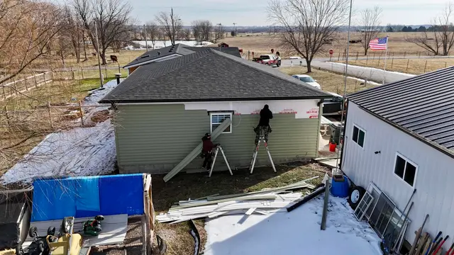Crew replacing siding on a green house in winter, with a metal-roofed shed beside it